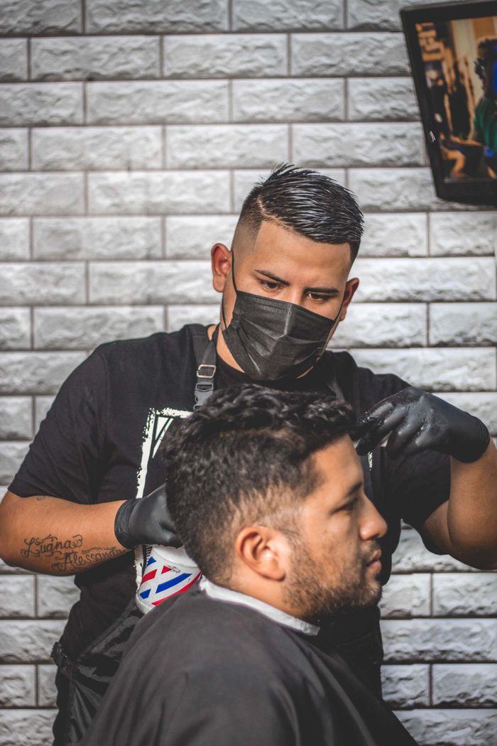 Barber wearing mask and gloves while styling client's hair in a contemporary barbershop setting.